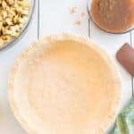 Unbaked pie crust in a glass pie dish with fork marks, surrounded by diced apples, melted butter, caramel sauce, and a green apple on a white surface.