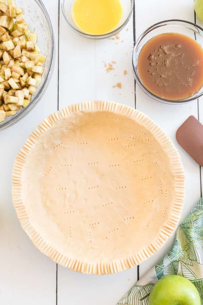 Unbaked pie crust in a glass pie dish with fork marks, surrounded by diced apples, melted butter, caramel sauce, and a green apple on a white surface.
