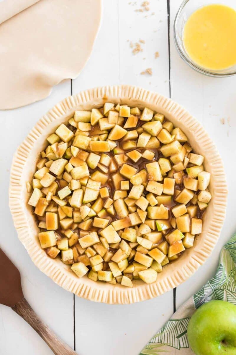Unbaked pie crust filled with chopped apples and brown sugar mixture on a white surface, with rolled dough, melted butter, and a green apple nearby.