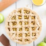 Unbaked apple pie with a decorative braided lattice crust, surrounded by apples, a rolling pin, a bowl of egg wash, and a spatula on a white surface.