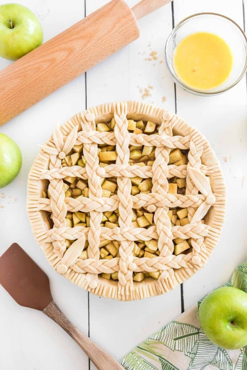 Unbaked apple pie with a decorative braided lattice crust, surrounded by apples, a rolling pin, a bowl of egg wash, and a spatula on a white surface.
