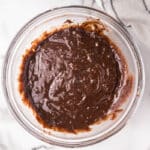 A clear glass bowl filled with chocolate brownie batter sits on a white marble surface next to a striped kitchen towel.