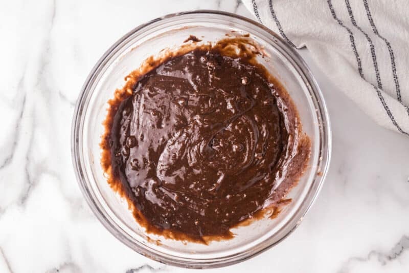 A clear glass bowl filled with chocolate brownie batter sits on a white marble surface next to a striped kitchen towel.