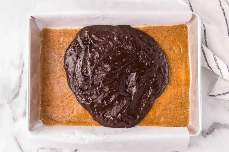 A rectangular baking pan with a baked cookie crust and chocolate batter spread on top, ready to be layered.
