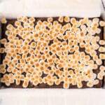 A rectangular baking pan lined with parchment paper contains a layer of toasted mini marshmallows on top of a chocolate base.