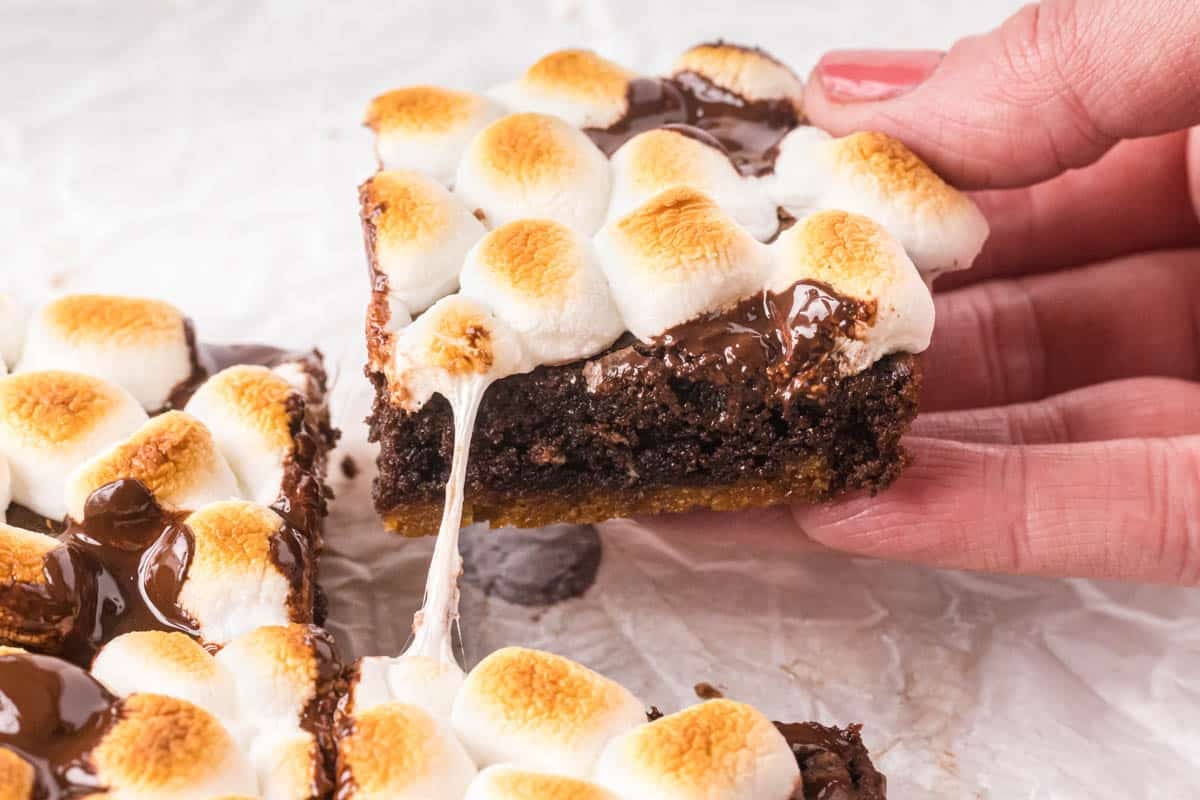 A hand holds a s'mores brownie topped with toasted marshmallows, chocolate, and a graham cracker crust, with melted marshmallow stretching from the bar.