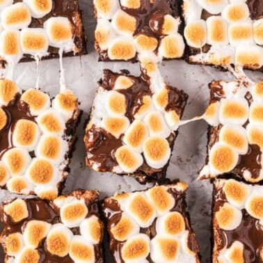 Brownies topped with toasted marshmallows and melted chocolate, cut into squares and arranged on parchment paper with pieces of graham crackers and mini marshmallows nearby.
