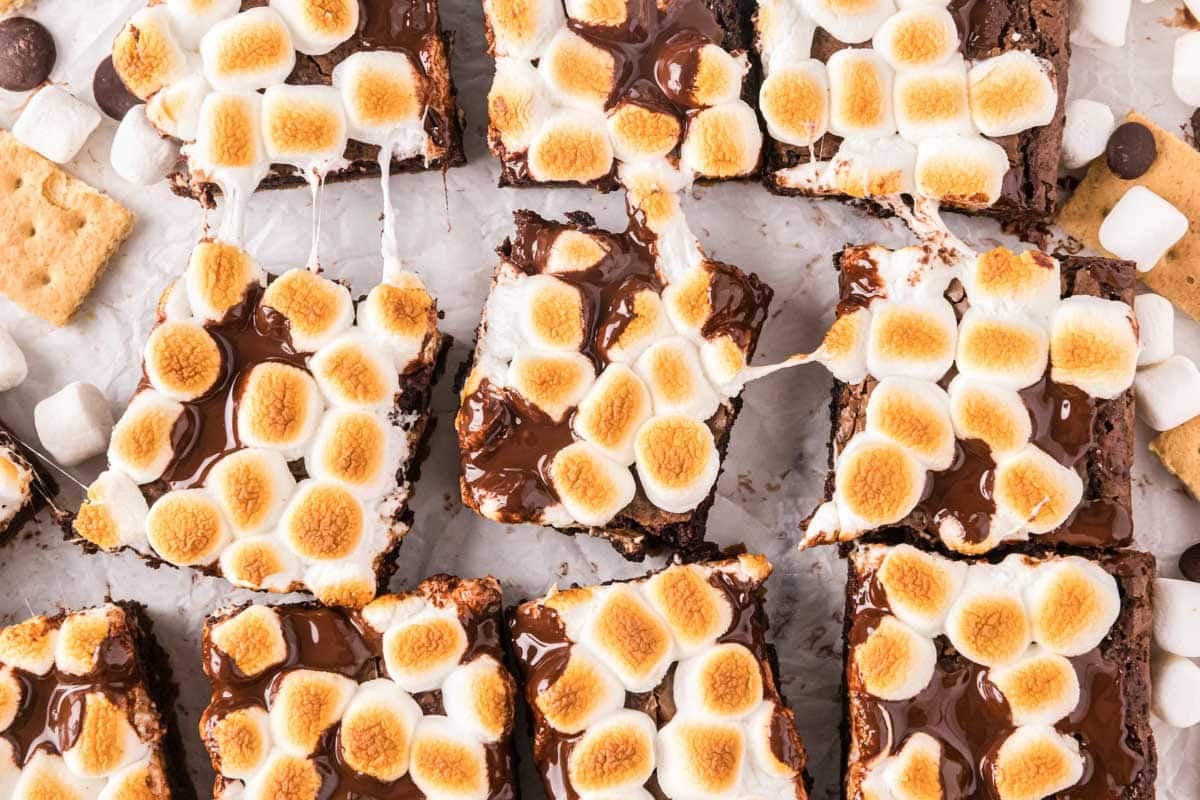 Brownies topped with toasted marshmallows and melted chocolate, cut into squares and arranged on parchment paper with pieces of graham crackers and mini marshmallows nearby.