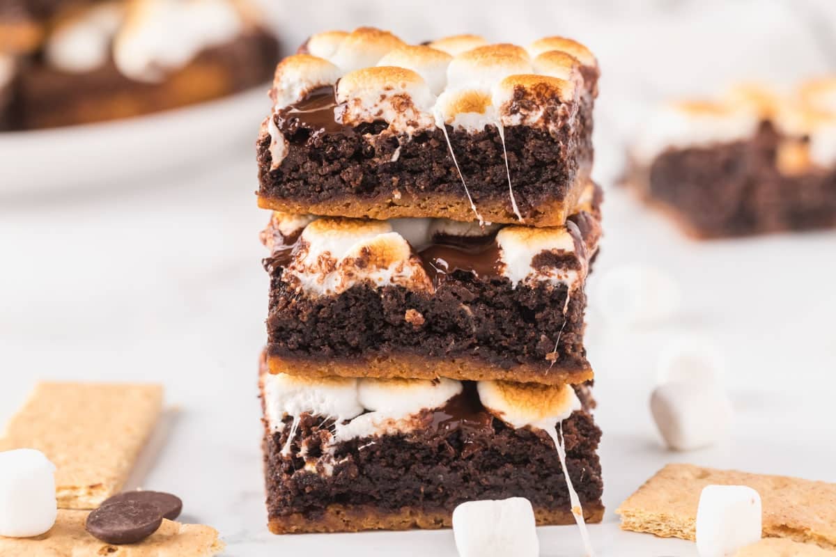 Three stacked s'mores brownies with toasted marshmallows and melted chocolate on top, surrounded by graham crackers and marshmallows.