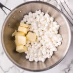 A stainless steel saucepan contains cubed butter and mini marshmallows, placed on a marble countertop next to a white and gray striped kitchen towel.