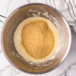 A metal mixing bowl contains a mixture of melted butter and graham cracker crumbs, next to a white kitchen towel with black stripes on a marble surface.