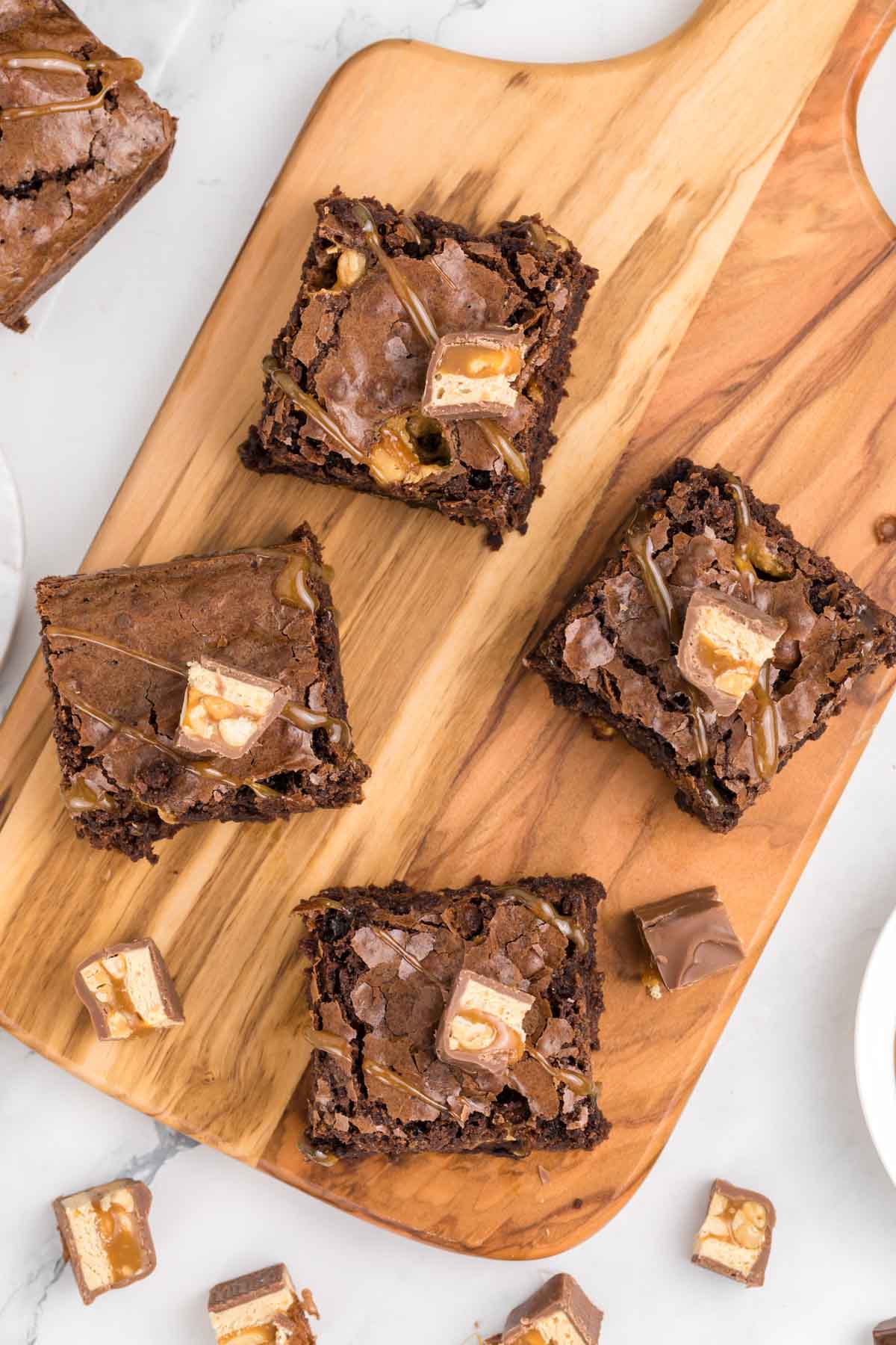 Four snickers brownies topped with pieces of candy bars are arranged on a wooden board, with additional candy pieces scattered around on a white surface.