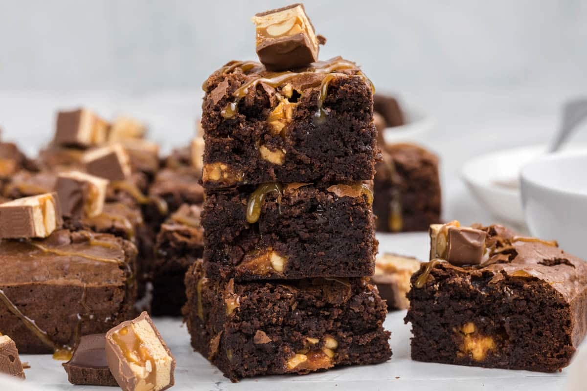 A stack of snickers brownies with caramel and candy pieces, surrounded by more brownies and candy chunks on a white surface.