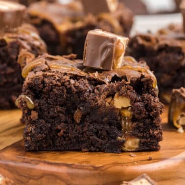 A close-up of a chocolate brownie with snickers with caramel and nut pieces, topped with a small chocolate candy, on a wooden board.