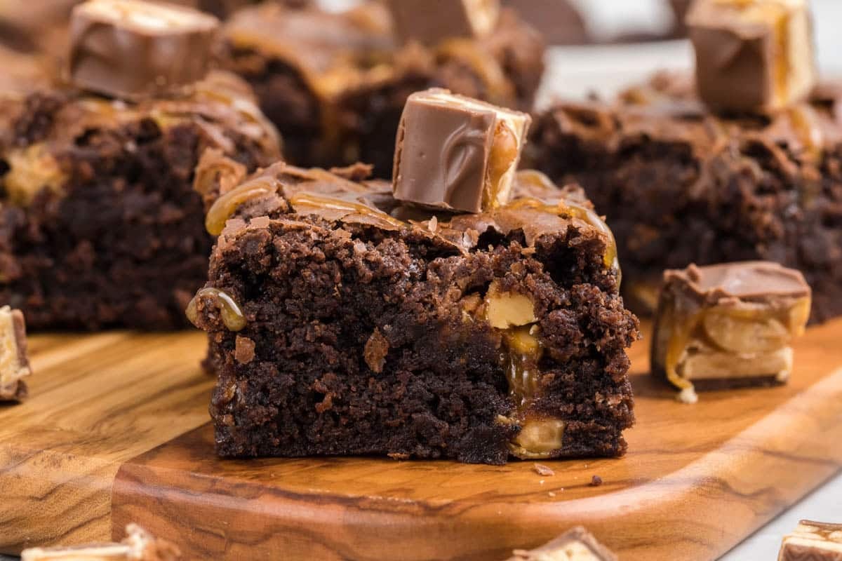 A close-up of a chocolate brownie with snickers with caramel and nut pieces, topped with a small chocolate candy, on a wooden board.