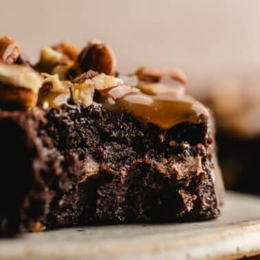 Close-up of a chocolate turtle brownie topped with caramel, chopped pecans, and a sprinkle of sea salt on a ceramic plate.