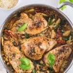 A skillet filled with cooked chicken breasts, spinach, sun-dried tomatoes, and artichoke hearts, garnished with fresh basil leaves. Parmesan and capers are in small bowls nearby.