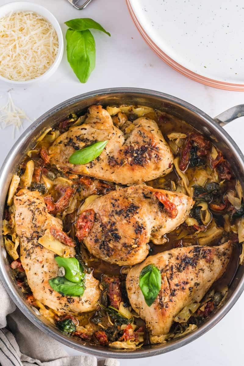 A stainless steel pan with cooked tuscan chicken breasts, sun-dried tomatoes, artichokes, spinach, and fresh basil garnish sits on a white surface next to a bowl of grated cheese and stacked plates.