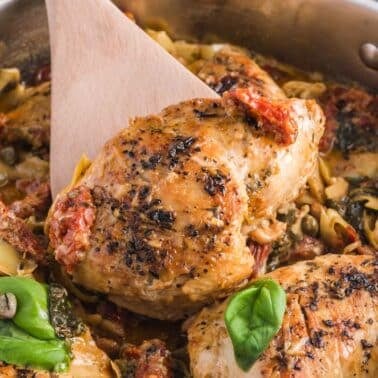 Cooked seasoned chicken breasts in a skillet with vegetables, herbs, sun-dried tomatoes, and a wooden spatula.