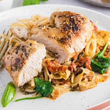 Sliced seasoned chicken breast served on a bed of pasta with spinach, sun-dried tomatoes, and creamy sauce, garnished with a basil leaf.