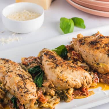 Three pieces of seasoned, cooked chicken breast served on a white platter with a mixture of vegetables and sauce, with a bowl of grated cheese and plates in the background.