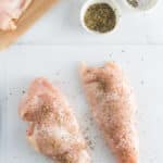 Two raw chicken breasts on a white cutting board, sprinkled with herbs and seasoning; more raw chicken and small bowls of herbs and salt are nearby.