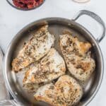 Four seasoned chicken breasts cooking in a stainless steel pan, with bowls of sun-dried tomatoes and minced garlic nearby on a white countertop.