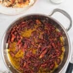A skillet filled with sun-dried tomatoes in oil sits on a white surface next to a plate of cooked chicken breasts and a glass of milk.