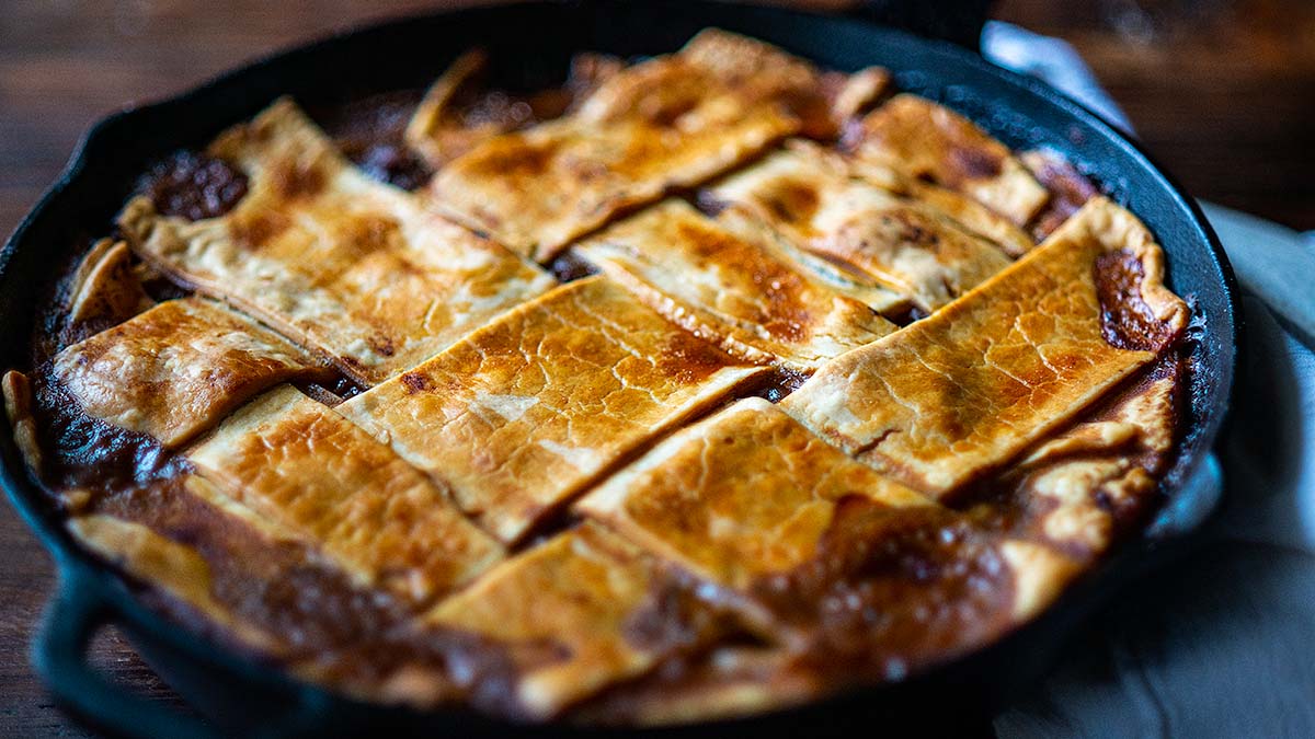 A skillet pie with a golden-brown lattice crust, packed with delicious pie fillings, is baked in a black cast iron pan and rests on a wooden surface.