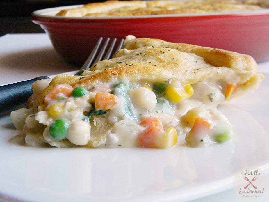 A slice of vegetable pot pie with a creamy pie filling and golden crust on a white plate, with a fork and red baking dish in the background—a classic among savory pies.