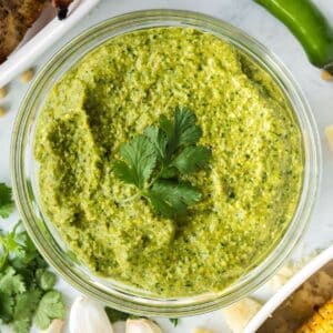 A glass bowl filled with green jalapeno pesto sauce, garnished with a sprig of fresh cilantro.