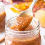 A spoon holding a thick, brown apple butter mixture over an open glass jar, with another jar and apples blurred in the background.