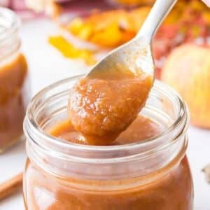 A spoon holding a thick, brown apple butter mixture over an open glass jar, with another jar and apples blurred in the background.