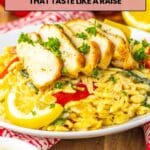 A plate of creamy pasta topped with sliced grilled chicken, garnished with parsley and lemon wedges, sits on a table with a red and white napkin.
