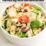 A white bowl filled with orzo pasta salad containing spinach, cherry tomatoes, cucumber, olives, red onion, and feta cheese, set on a white surface.