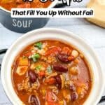 A bowl of hearty bean and vegetable soup sits on a white surface, with another bowl of soup and a piece of bread in the background. Text reads "16 Soups That Fill You Up Without Fail.