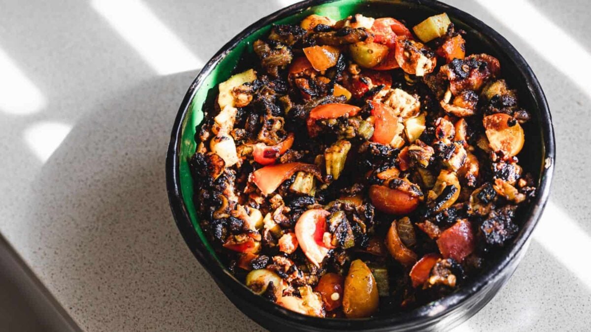 A green and black bowl filled with charred and roasted vegetables, including tomatoes and zucchini, sits on a light countertop—perfect inspiration for vegan recipes.