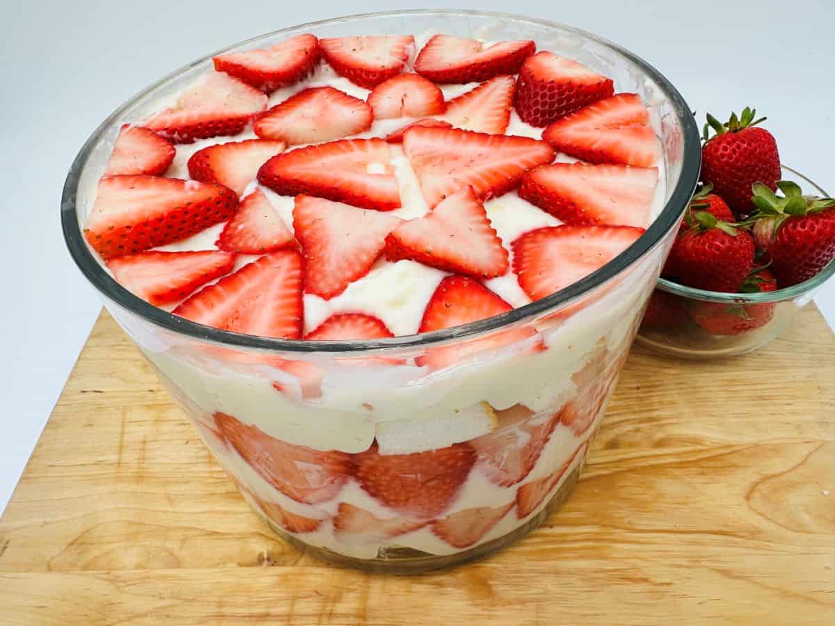A glass bowl filled with layered strawberry trifle—sliced strawberries, cream, and cake—alongside a small bowl of whole strawberries on a wooden surface. This is one of the perfect nut free desserts for those seeking allergy friendly snacks.