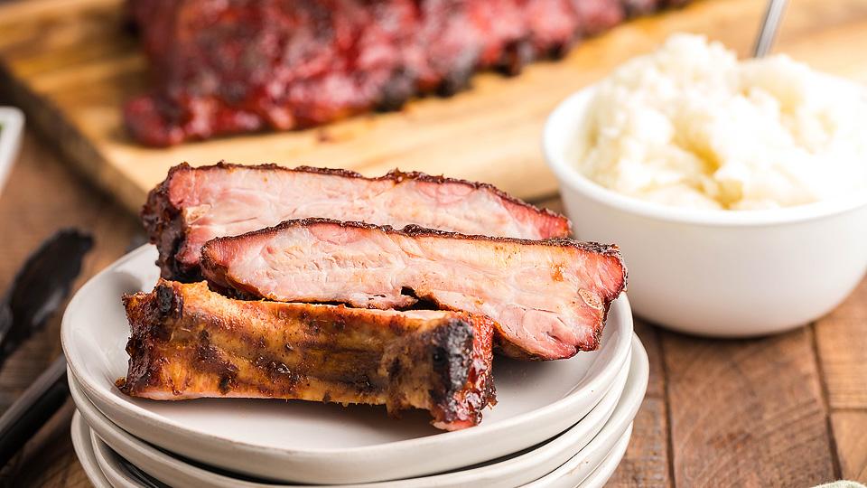 Three pieces of grilled ribs are stacked on a plate, with a bowl of mashed potatoes and more ribs on a wooden board in the background—a perfect choice for all-season recipes.