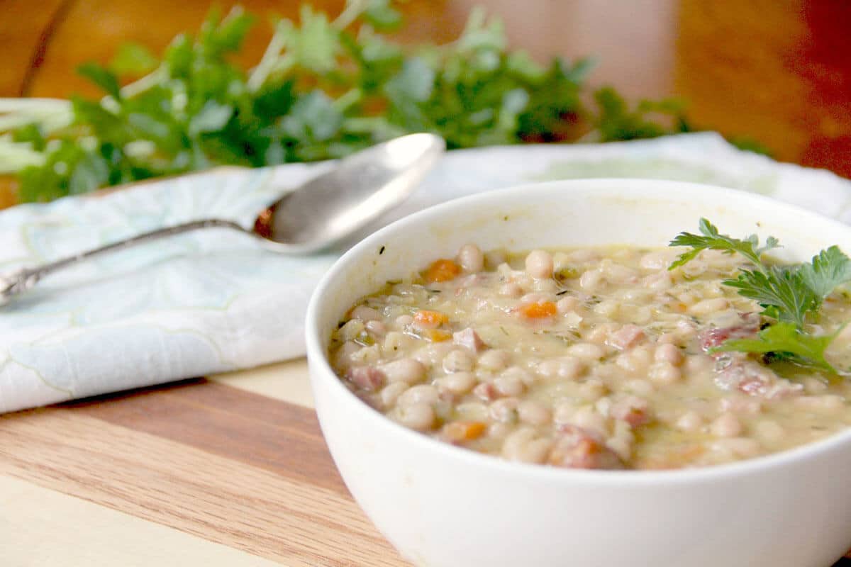 A white bowl filled with bean and vegetable soup sits on a wooden surface, garnished with parsley. Perfect for cozy recipes, a spoon and fresh herbs are in the background.