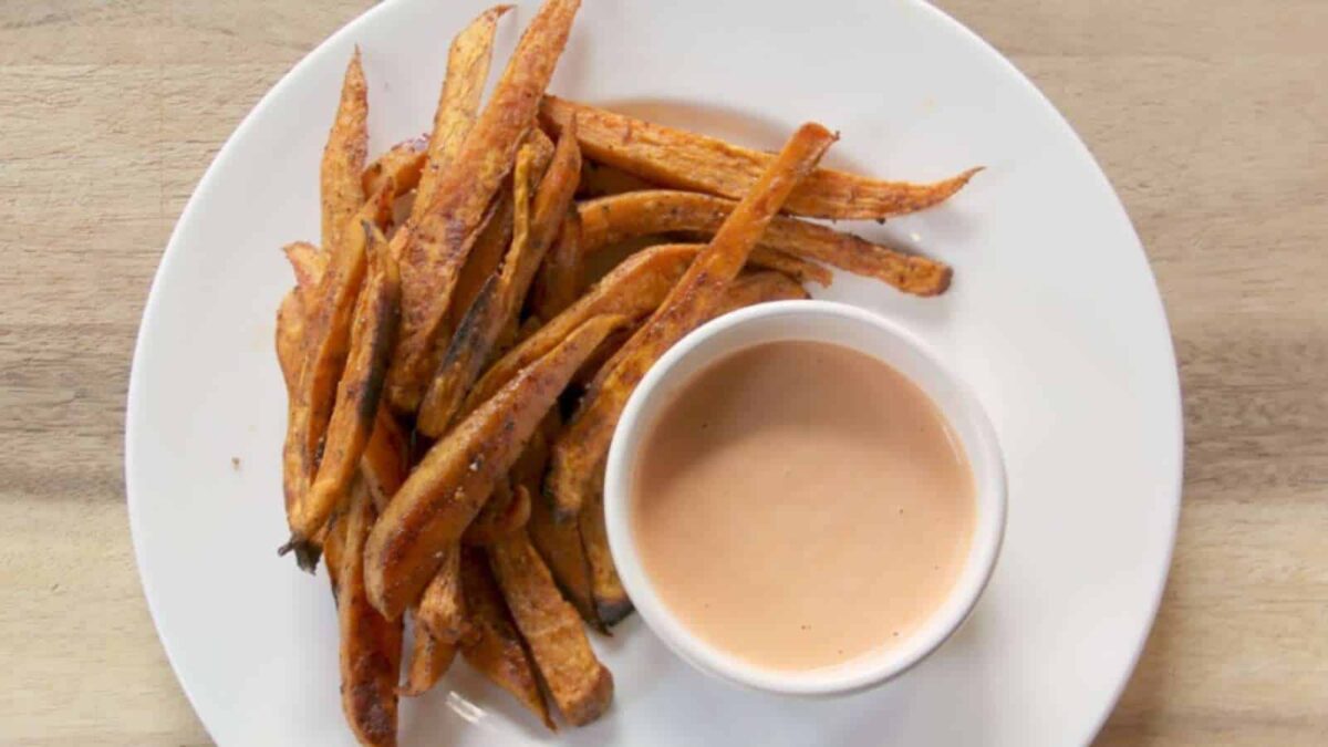 A white plate with Sweet Potato fries, served as one of our delicious Sides or Mains, accompanied by a small cup of light orange dipping sauce.