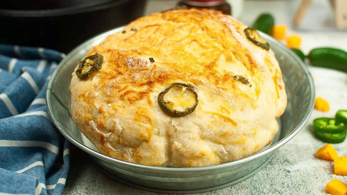 A round loaf of bread topped with sliced jalapeños and cheese sits in a metal baking pan on a light surface, perfect to bake and share for your next Holiday Breads spread.