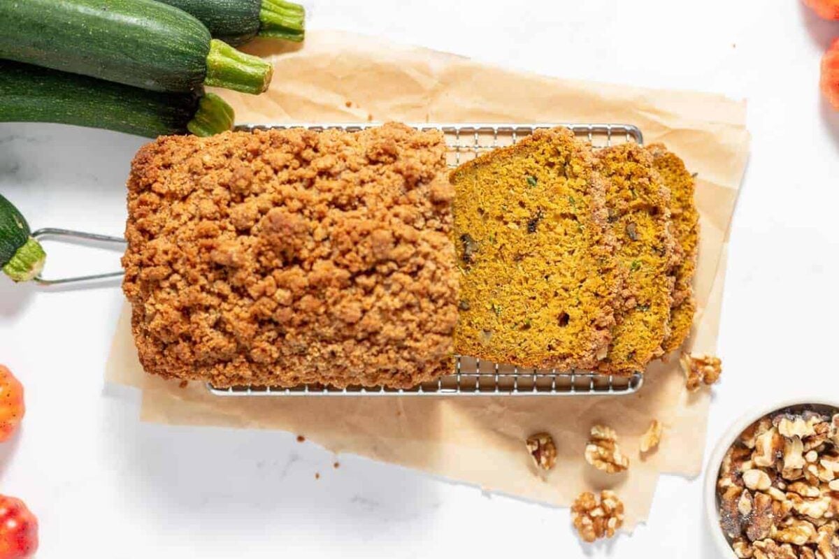 A loaf of zucchini bread with a crumb topping is partially sliced on a wire rack, perfect to share during the holidays, with zucchinis, walnuts, and apples nearby.