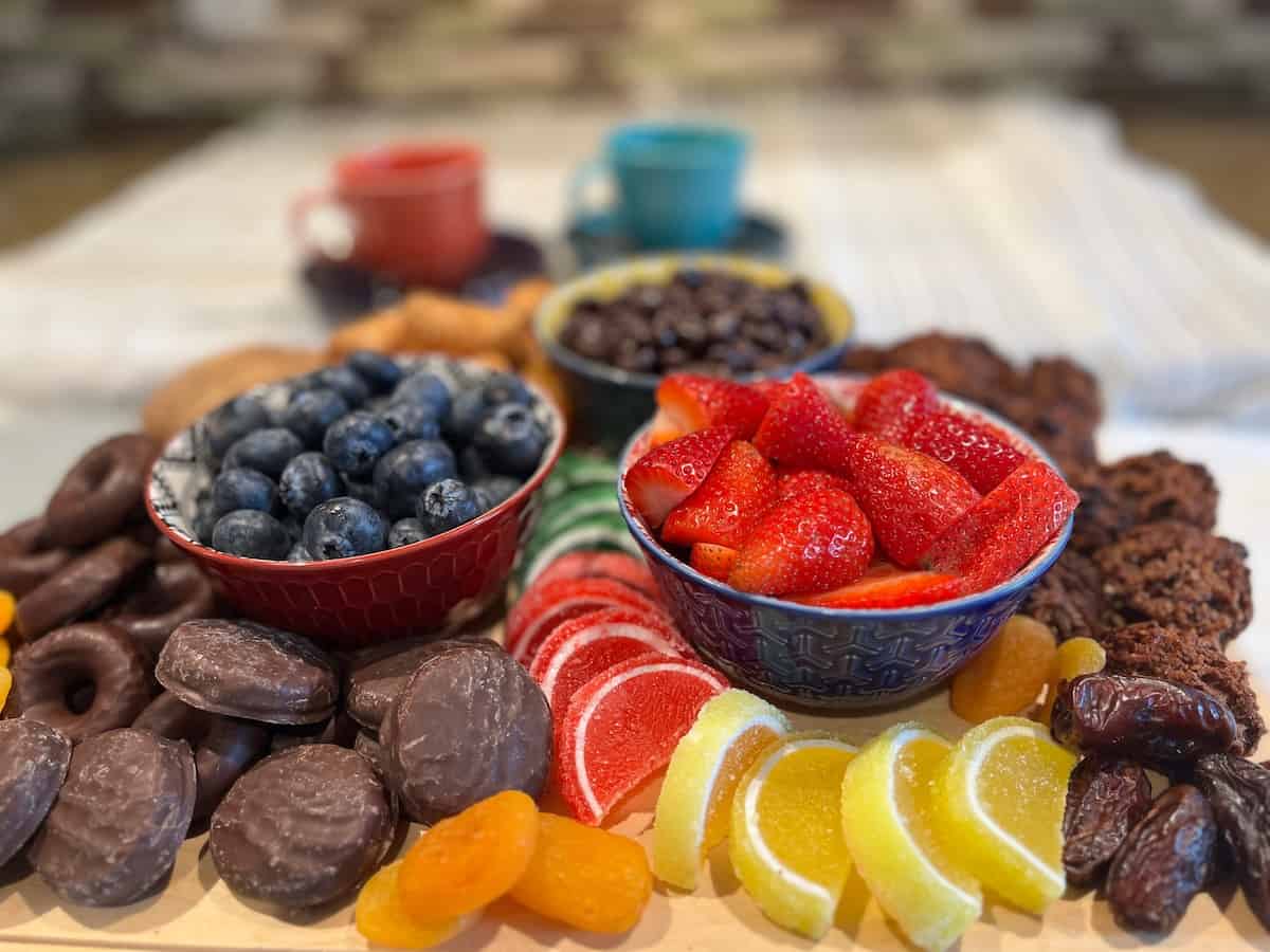A platter with bowls of blueberries and strawberries, surrounded by chocolate-covered treats, dried fruit, citrus jellies, dates, and three colorful cups in the background—perfect for entertaining or as party appetizers inspired by creative charcuterie ideas.