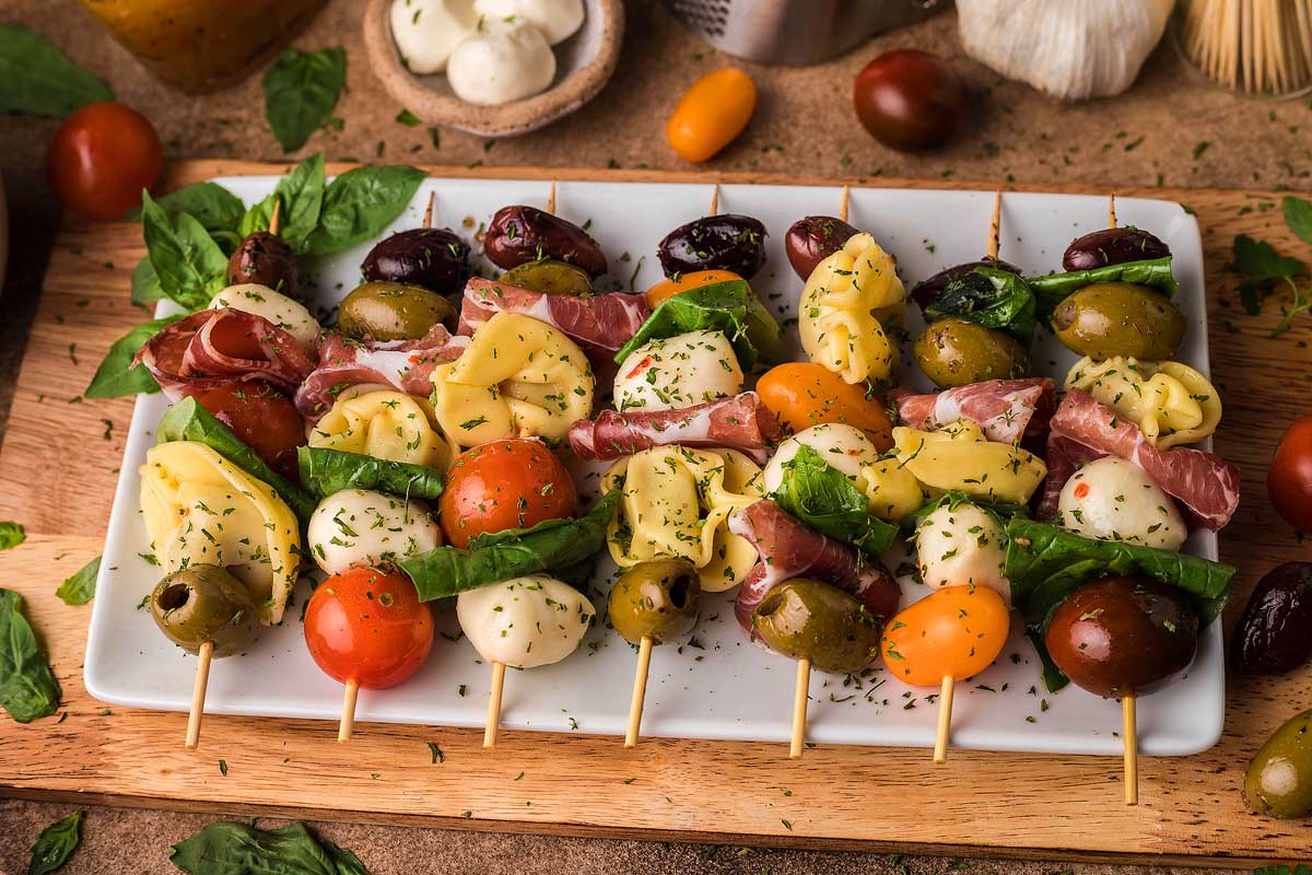A rectangular plate holds skewers with mozzarella balls, olives, cherry tomatoes, prosciutto, tortellini, and basil leaves, garnished with herbs—perfect for party appetizers or creative charcuterie ideas.