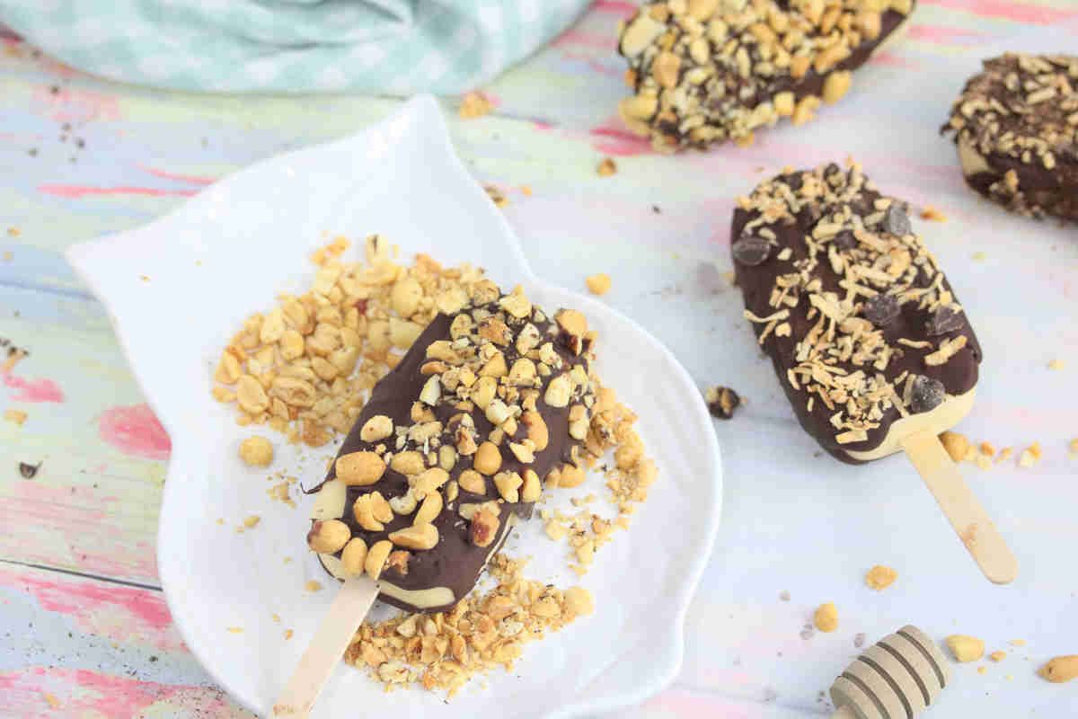 Two chocolate-covered ice cream bars on sticks, topped with chopped nuts, rest on a white plate with nut crumbs—a true comfort dessert—while more bars and crumbs add a cozy touch to the pastel-colored surface.