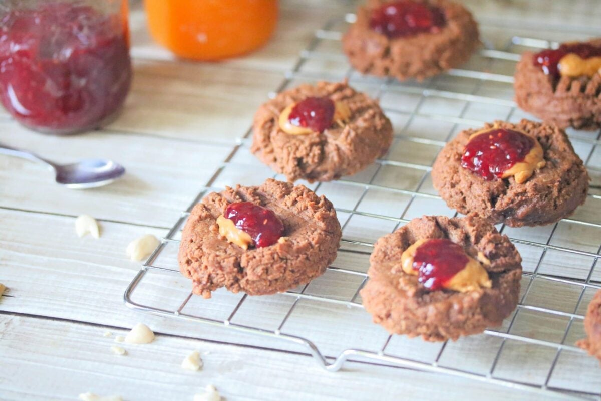 Chocolate cookies with peanut butter and jelly centers are cooling on a wire rack, offering the perfect comfort desserts, with jars of jam and a spoon in the background.