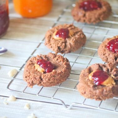 Chocolate cookies with peanut butter and jelly centers are cooling on a wire rack, offering the perfect comfort desserts, with jars of jam and a spoon in the background.