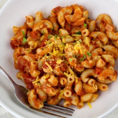 A white bowl filled with elbow macaroni in tomato sauce, topped with shredded cheese and herbs, showcases one of the classic pasta dishes, with a fork resting on the side.