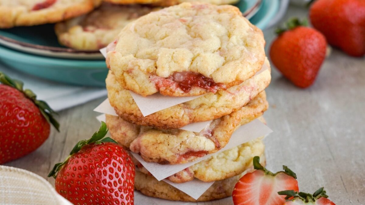 A stack of three easy cookies with strawberry flavor, separated by parchment paper, sits on a table, with fresh strawberries and more cookies in the background.